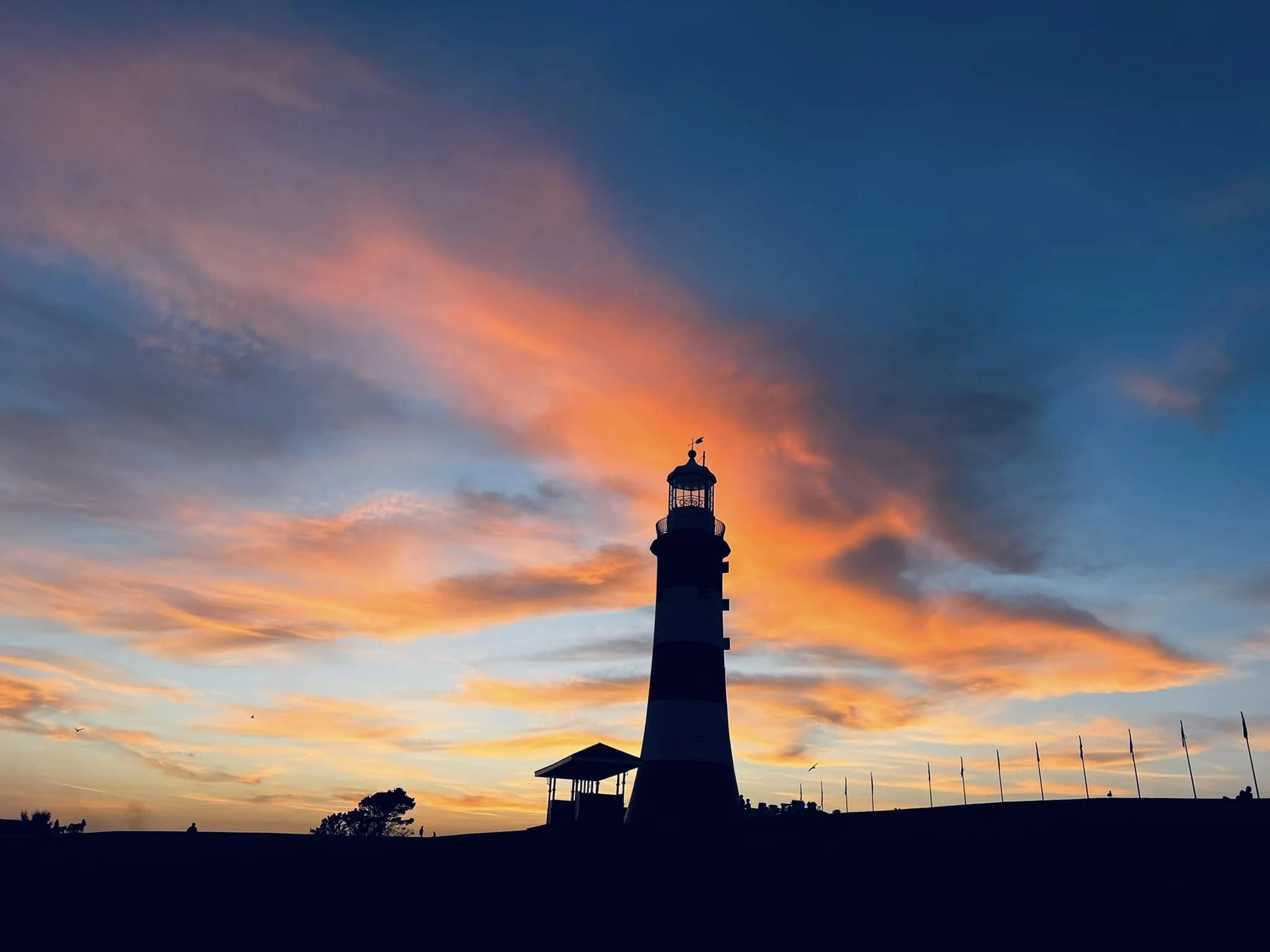 Smeaton's Tower lighthouse in silhouette against a fiery orange and blue sunset sky on Plymouth Hoe, with a small pavilion and a row of flagpoles along the horizon.